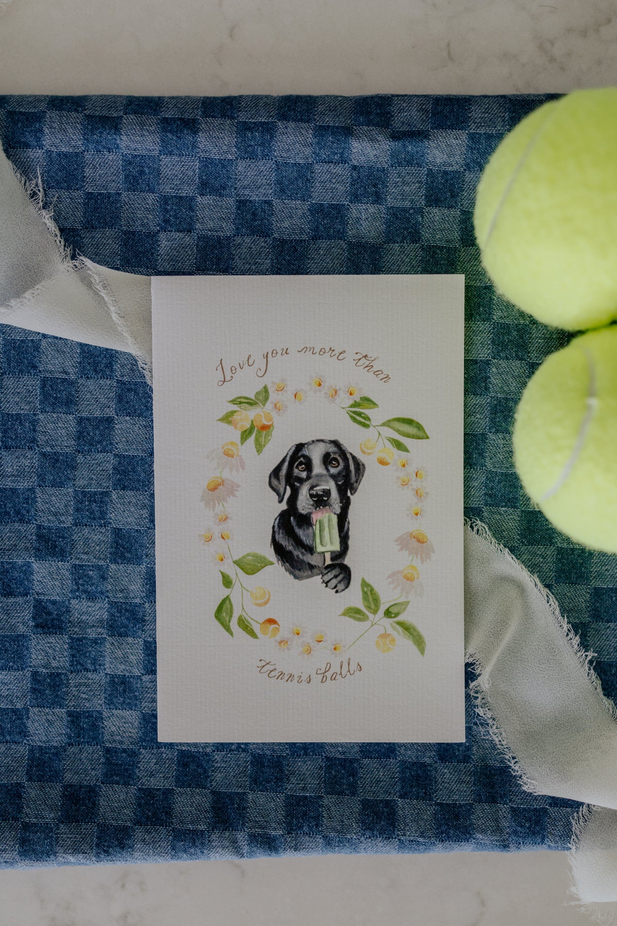 A square white greeting card featuring a watercolor illustration of a black lab surrounded by green leaves and yellow tennis balls, with the text 'Love you more than Tennis Balls'. The card is styled on a blue and white checkered fabric alongside two yellow tennis balls and white ribbon.