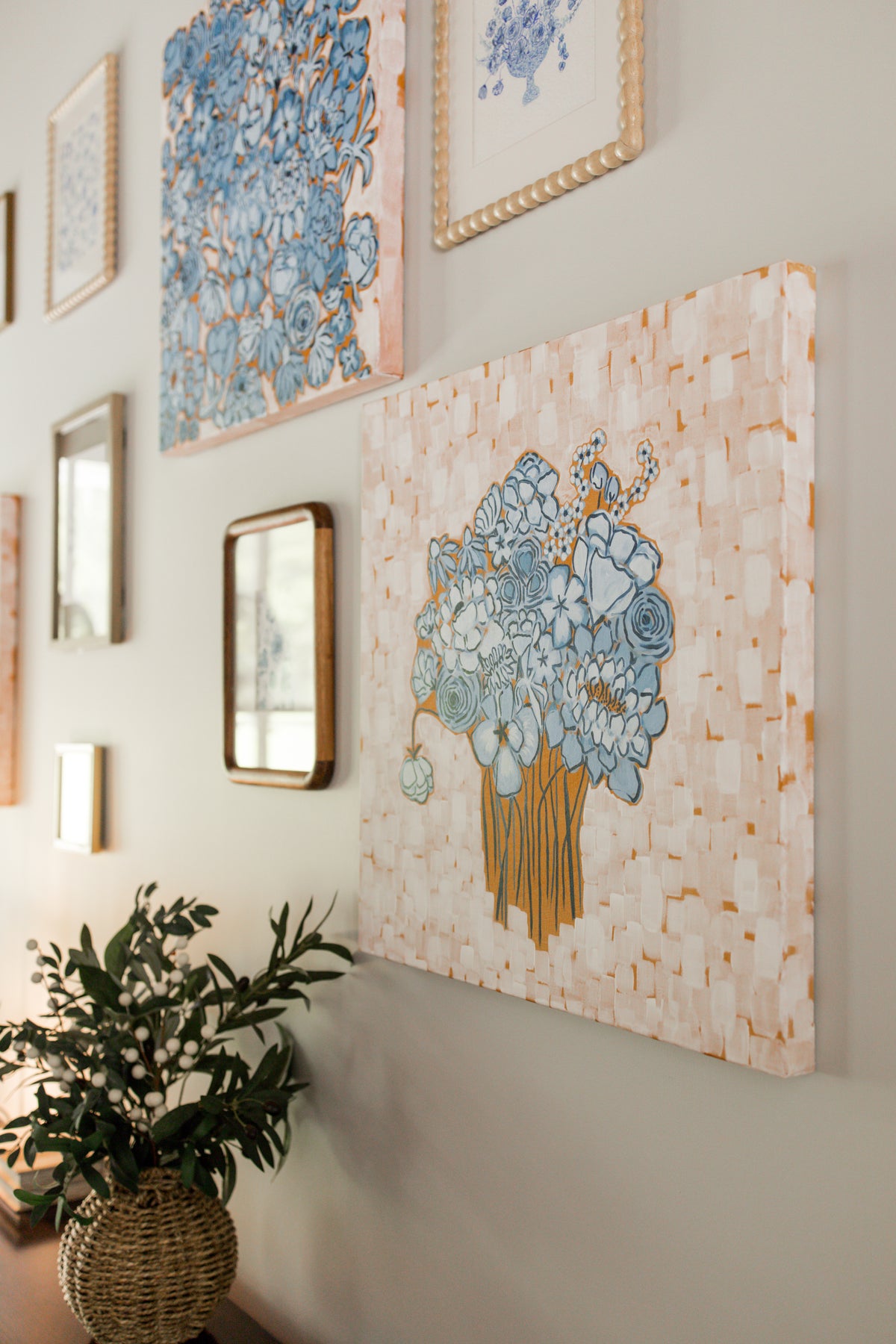 A large canvas wall art print featuring a bouquet of blue flowers in a vase, displayed on a beige wall alongside other framed art and a potted plant.