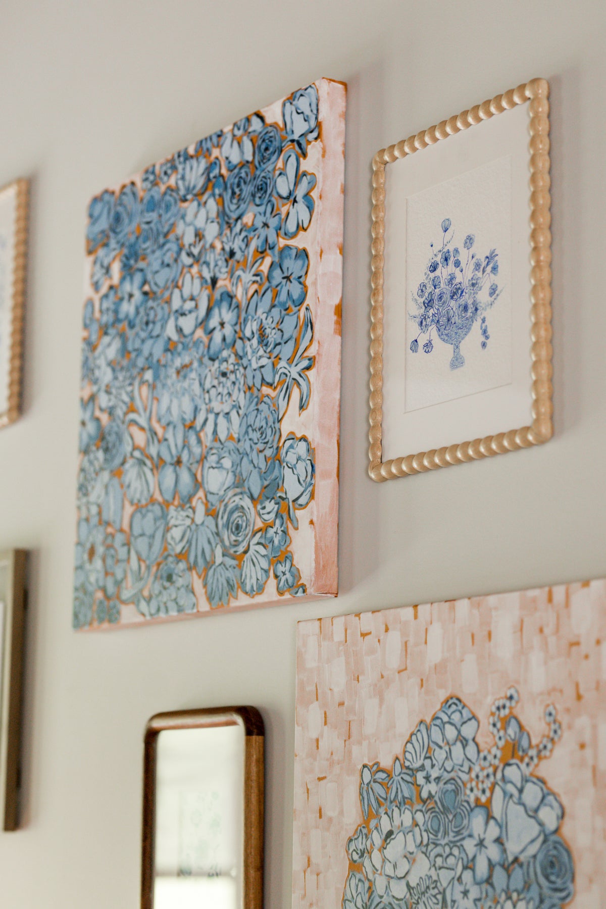 A gallery wall display featuring multiple framed art prints. The most prominent piece is a square canvas with a blue floral pattern outlined in a wood frame, hanging next to a smaller framed print with a blue floral bouquet and wooden beaded frame.