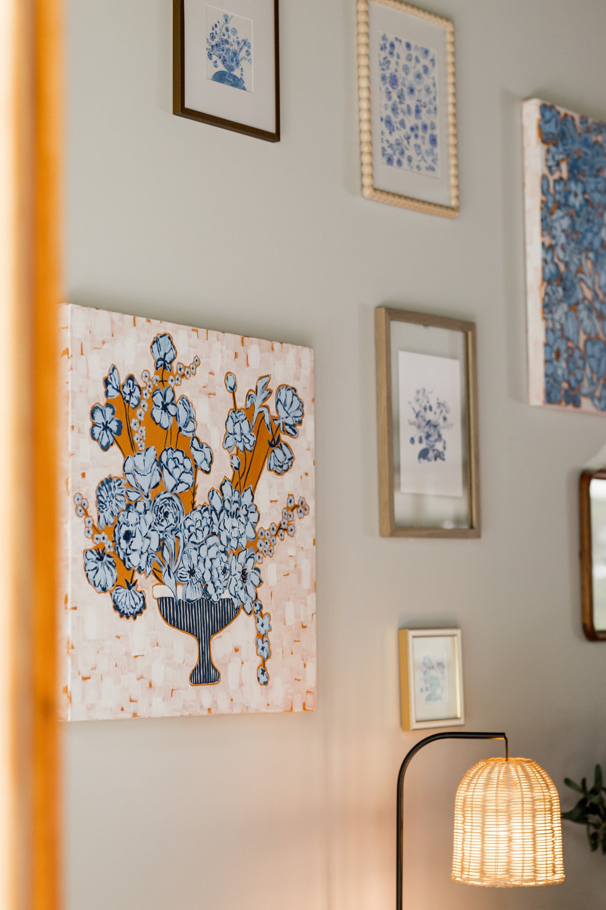 A framed floral wall art print with blue flowers and hanging on a light gray gallery wall, alongside other framed prints and a warm glowing woven lamp.