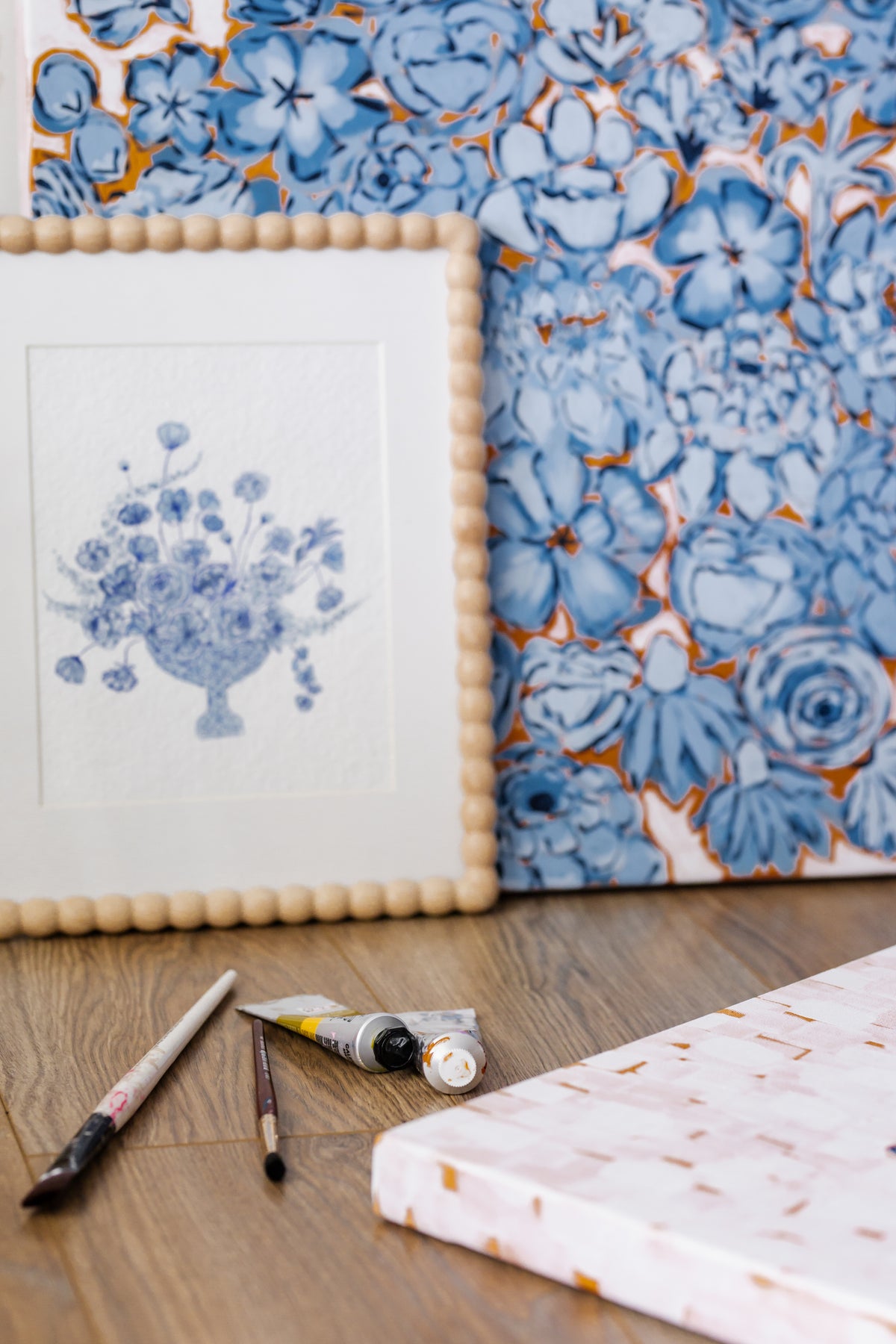 A styled flat-lay of art supplies on a wooden floor, featuring a framed watercolor painting of a floral bouquet in a blue and white palette, a canvas with a blue and orange floral pattern, a tube of paint, two paintbrushes, and a folded piece of patterned fabric.