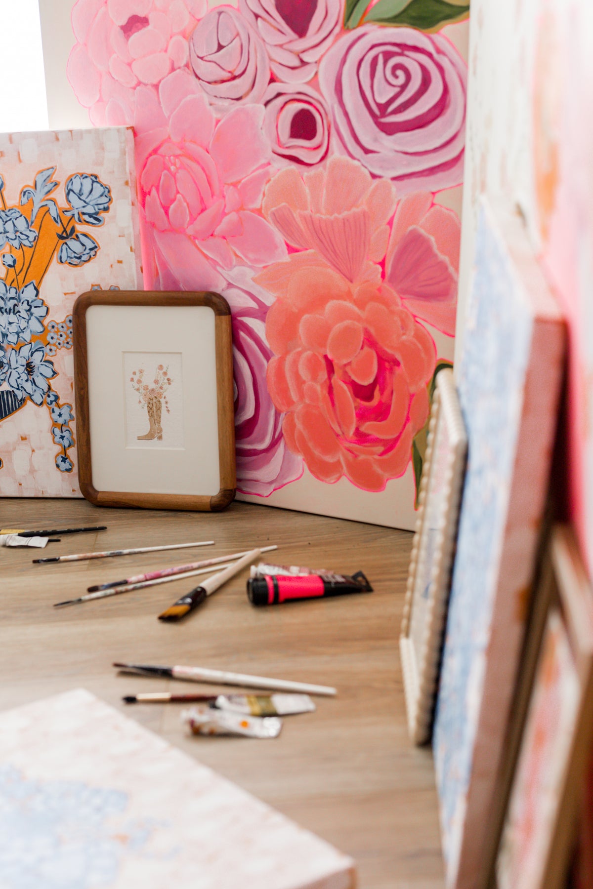 A styled art studio scene featuring a large floral canvas print with pink and coral roses, a small framed artwork, and various paintbrushes, tubes of paint, and other canvases on a wooden table.