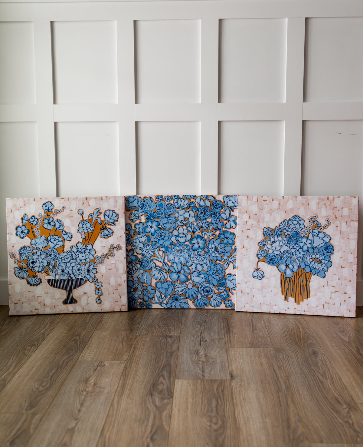 Three square decorative canvases with floral patterns in blue, orange, and white, leaning against a white paneled wall on a wood floor.