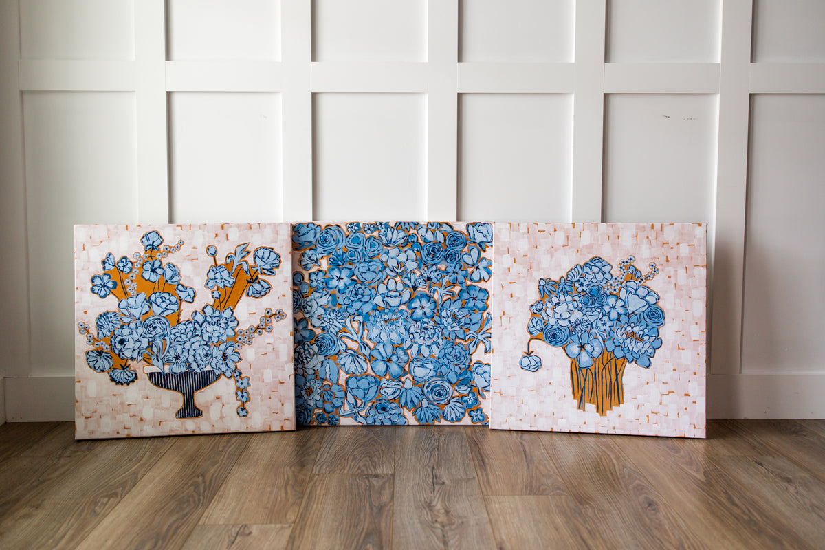 Three square decorative canvases with floral patterns in blue, orange, and white, leaning against a white paneled wall on a wood floor.