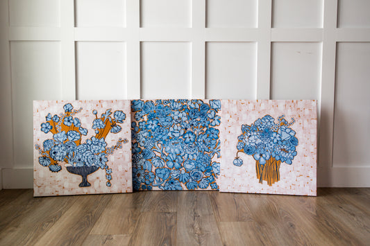 Three square decorative canvases with floral patterns in blue, orange, and white, leaning against a white paneled wall on a wood floor.