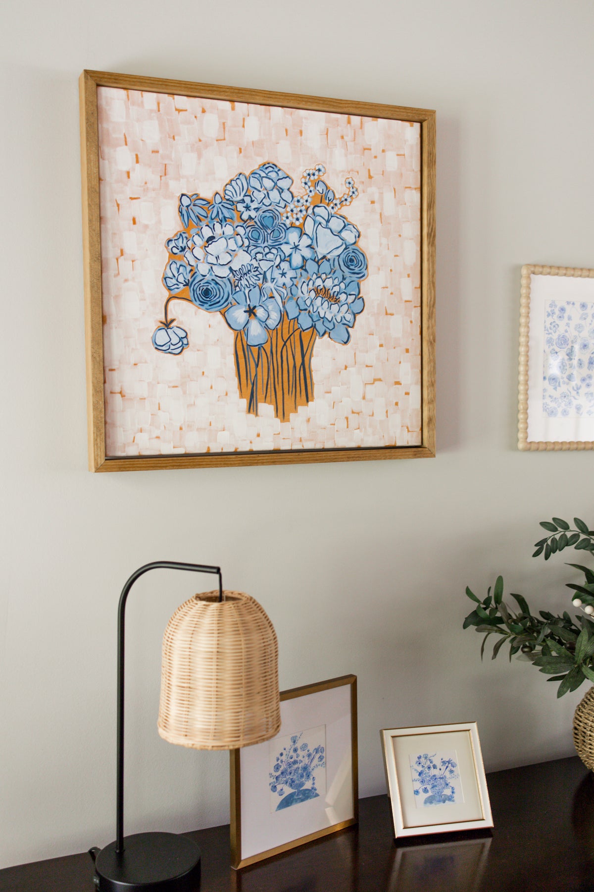 A framed floral art print featuring blue flowers with an orange background with a textured cream overlay, displayed on a light gray wall above a dark wooden table with a lamp and small framed prints.