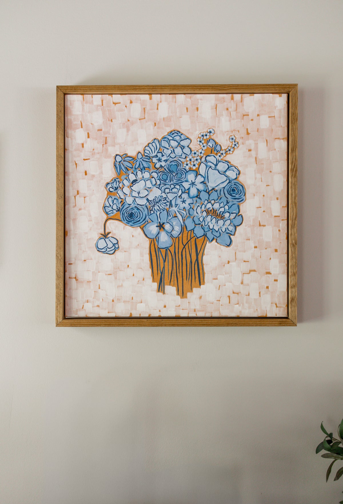 A framed floral art print featuring a bouquet of light blue flowers on a textured orange and white background, enclosed in a light wood frame and mounted on a neutral wall.