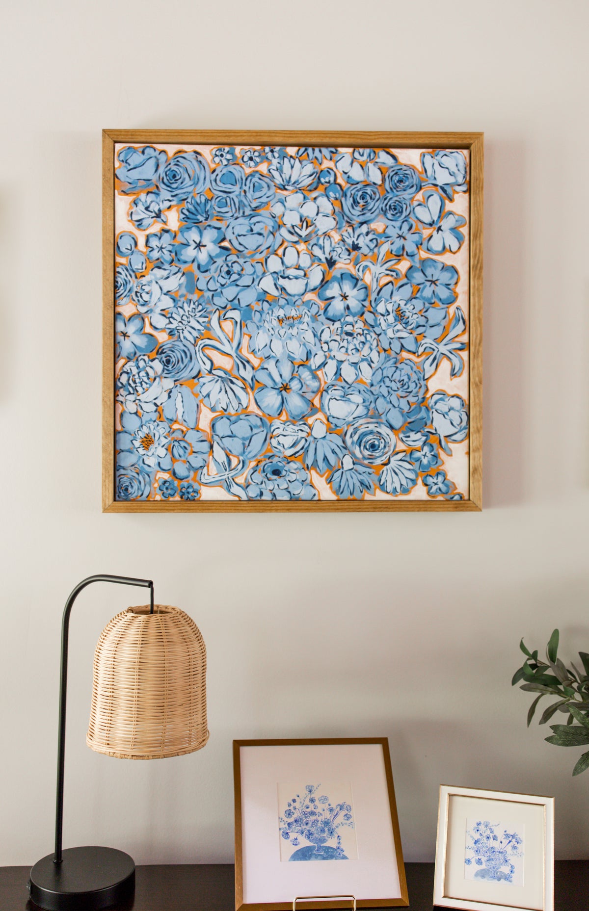 A framed floral canvas featuring blue flowers and orange background in a light wood frame, displayed on a wall above a console table with a lamp, small framed prints, and a potted plant.