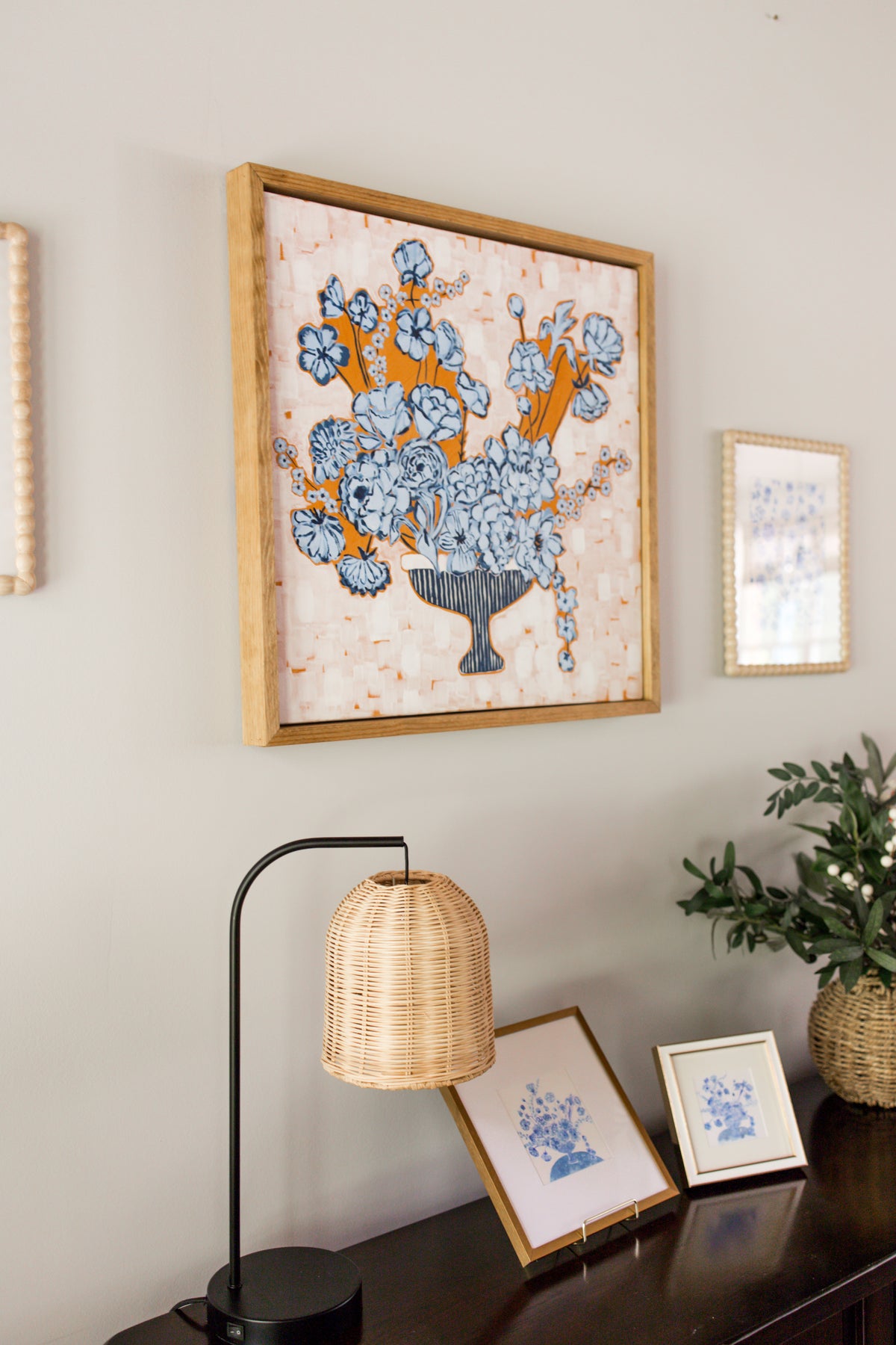 A framed floral canvas featuring blue flowers and orange background in a light wood frame, displayed on a wall above a console table with a lamp, small framed prints, and a potted plant.