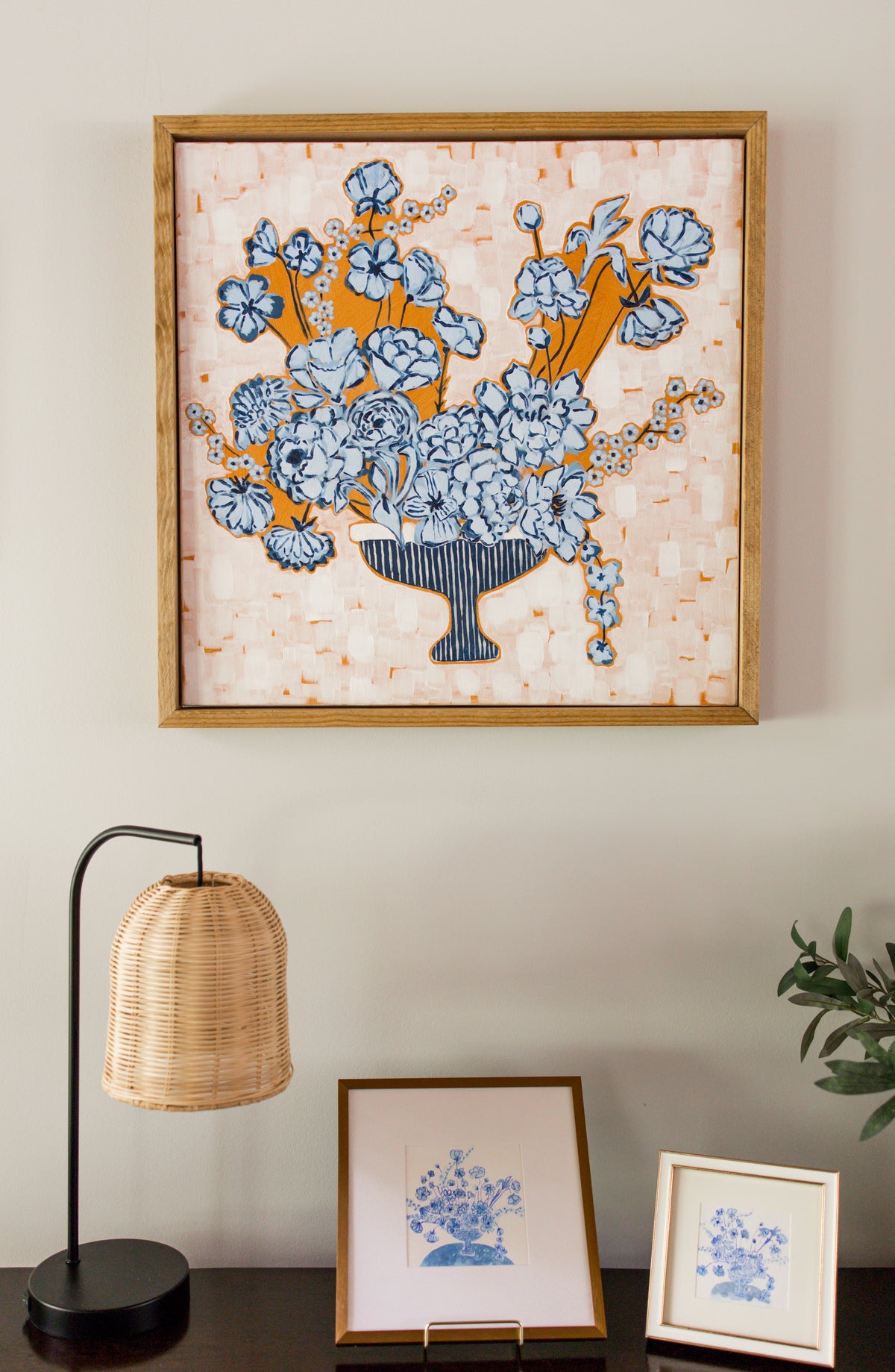 A framed canvas featuring a blue and orange floral bouquet on a textured cream background, hanging on a light gray wall above a dark table with a woven lamp and smaller framed prints.