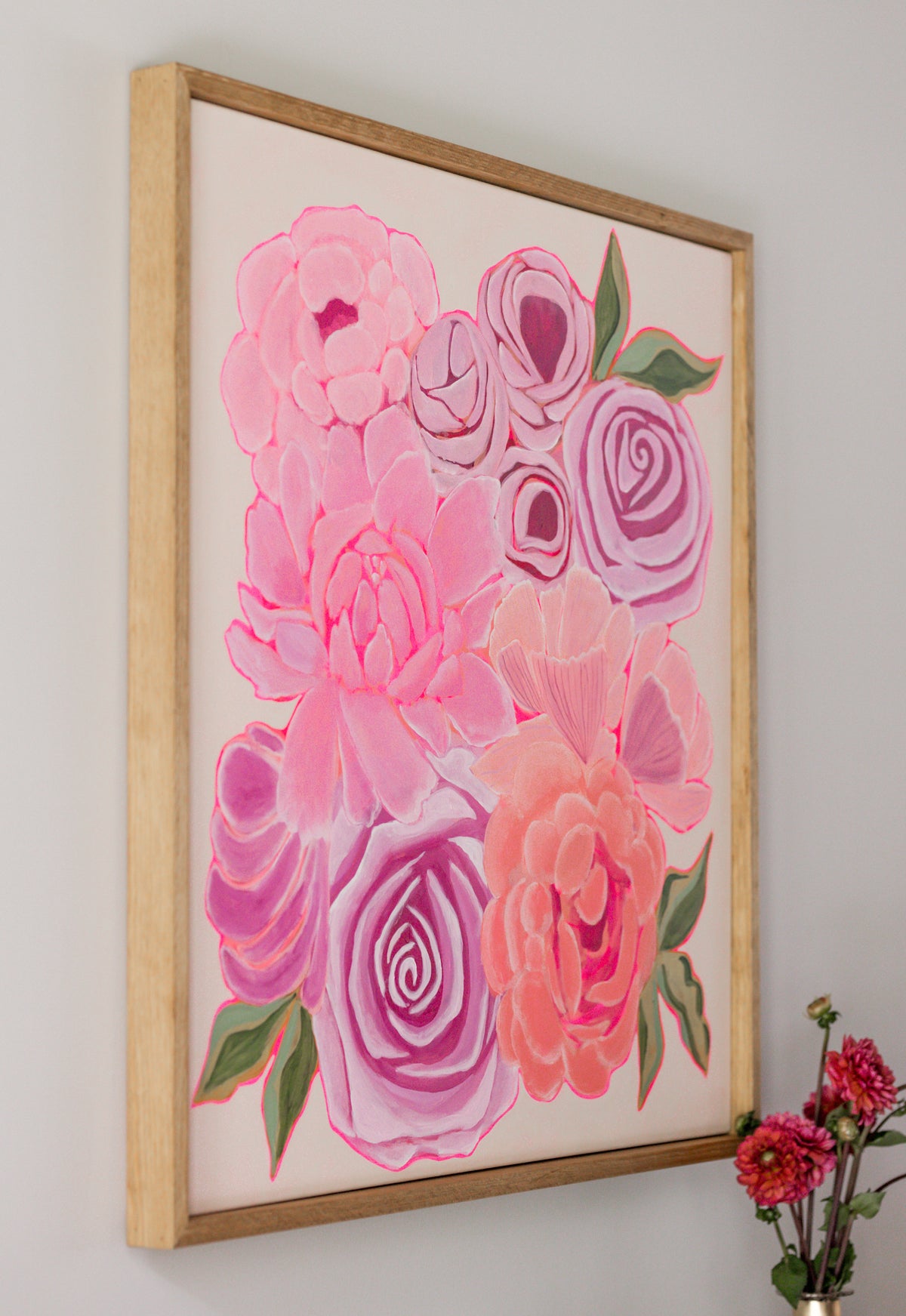 Framed wall art featuring a stylized floral painting with pink and coral roses, green leaves, and a light wood frame hanging on a white wall. A small vase with flowers appears in the lower right corner.