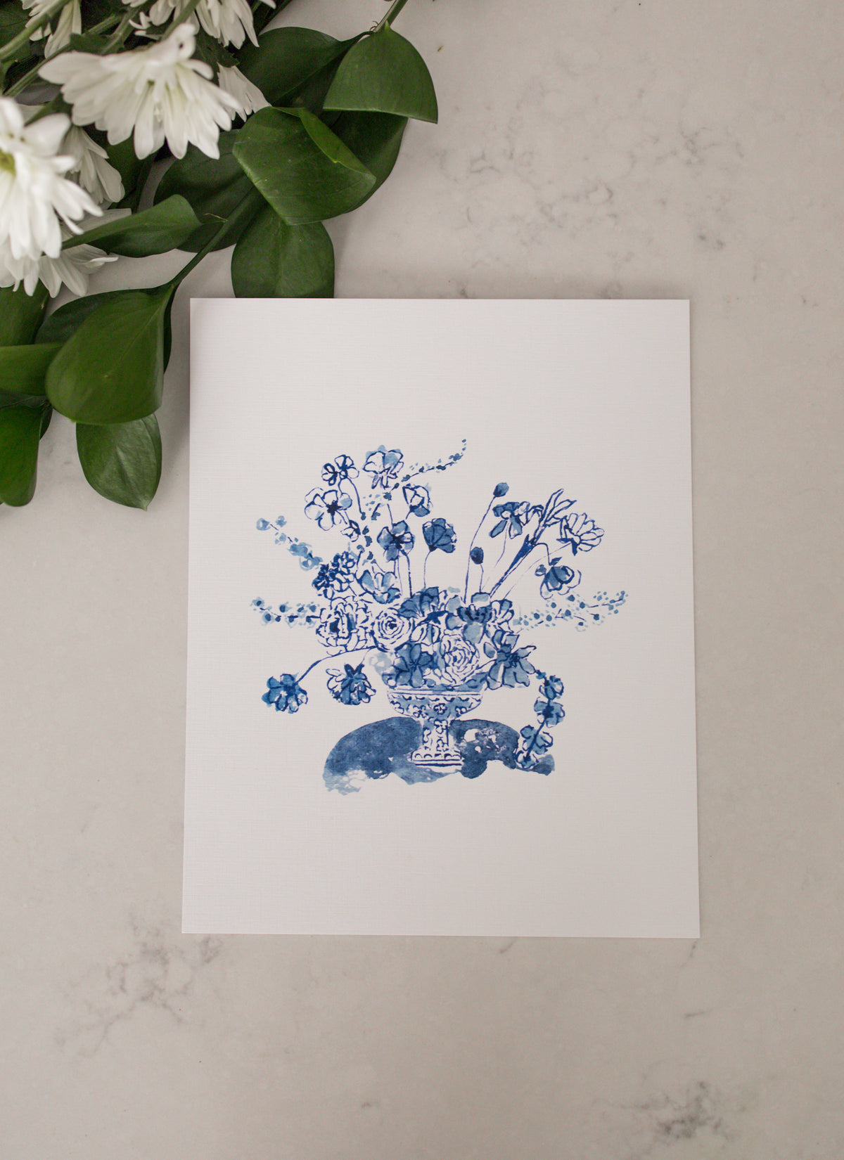 Rectangular art print of a Blue bouquet arrangement surrounded by white flowers with greenery on a marble surface 