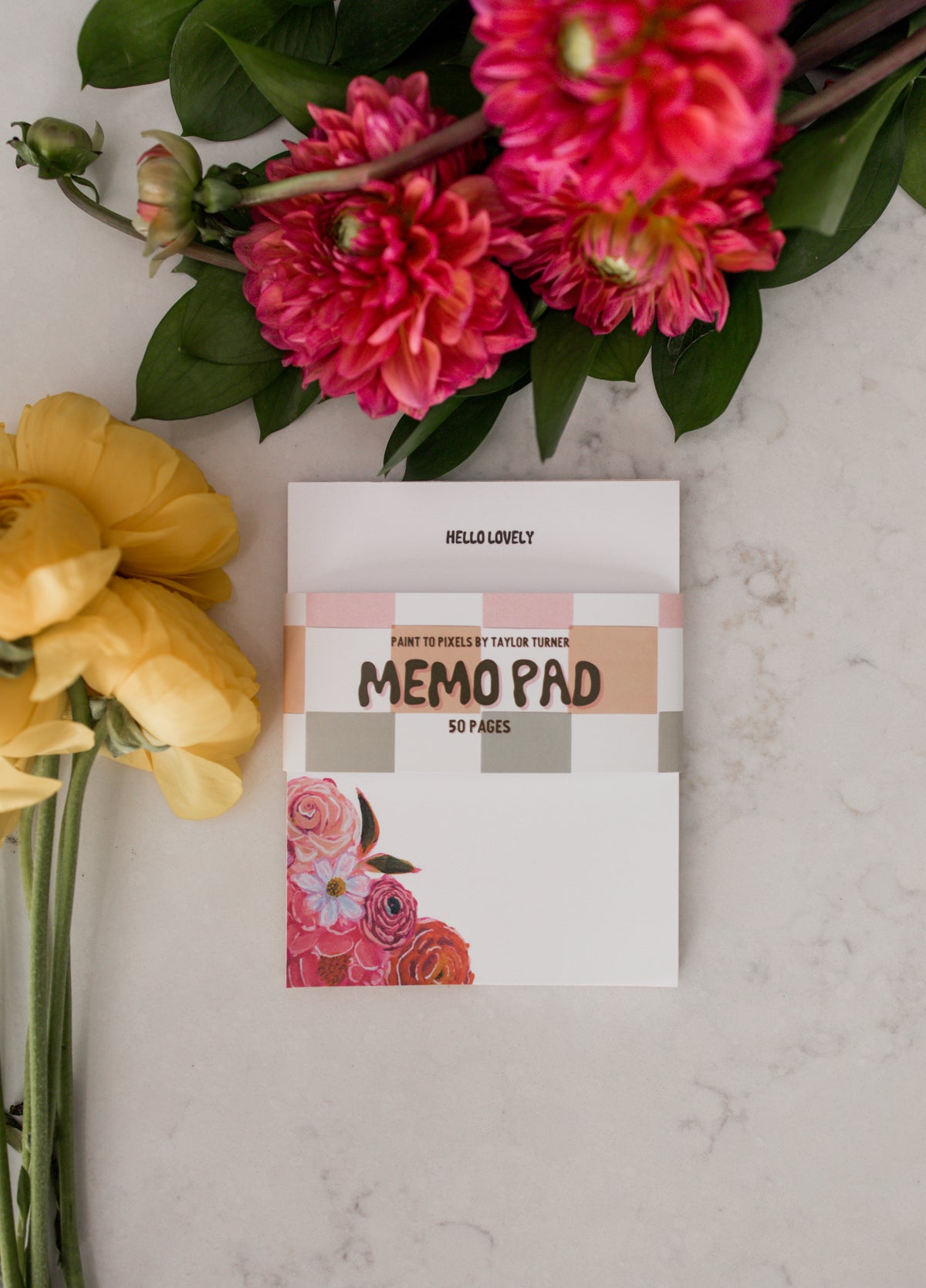 A memo pads styled on a light marble surface with yellow and pink flowers. The memo pad reads 'HELLO LOVELY' with a checkered belly band to describe the product that says "PAINT TO PIXELS BY TAYLOR TURER, MEMO PAD, 50 PAGES" 