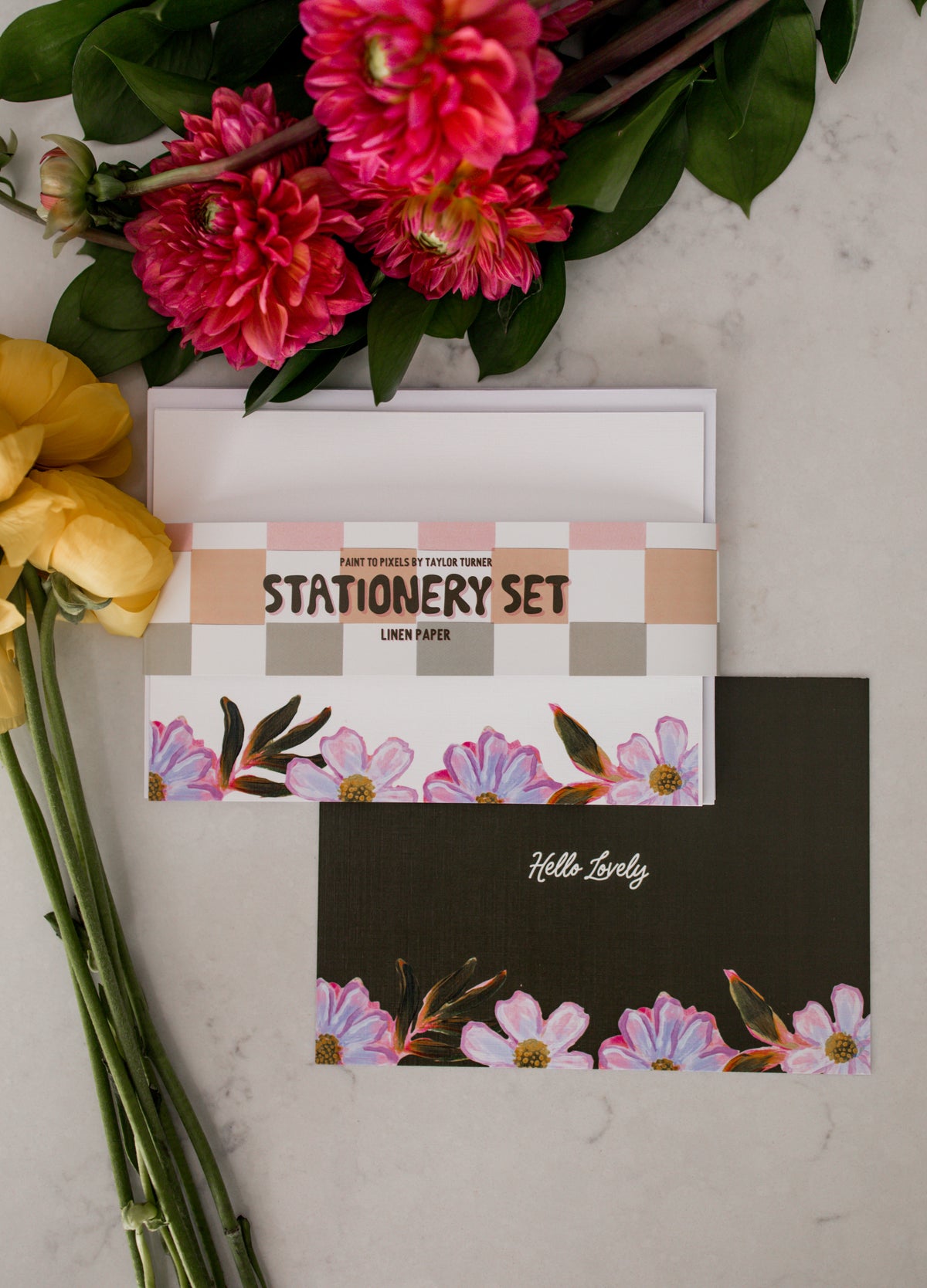 A stationery set displayed on a light marble surface with pink and yellow flowers. The backside of the card featured underneath is green and reads "Hello lovely" with brightly colored daisies at the bottom. On top is the front side of the stationery set which is white with brightly colored daisies at the bottom and a belly band holding the set together that reads "PAINT TO PIXELS BY TAYLOR TURNER, STATIONERY SET, LINEN PAPER." 