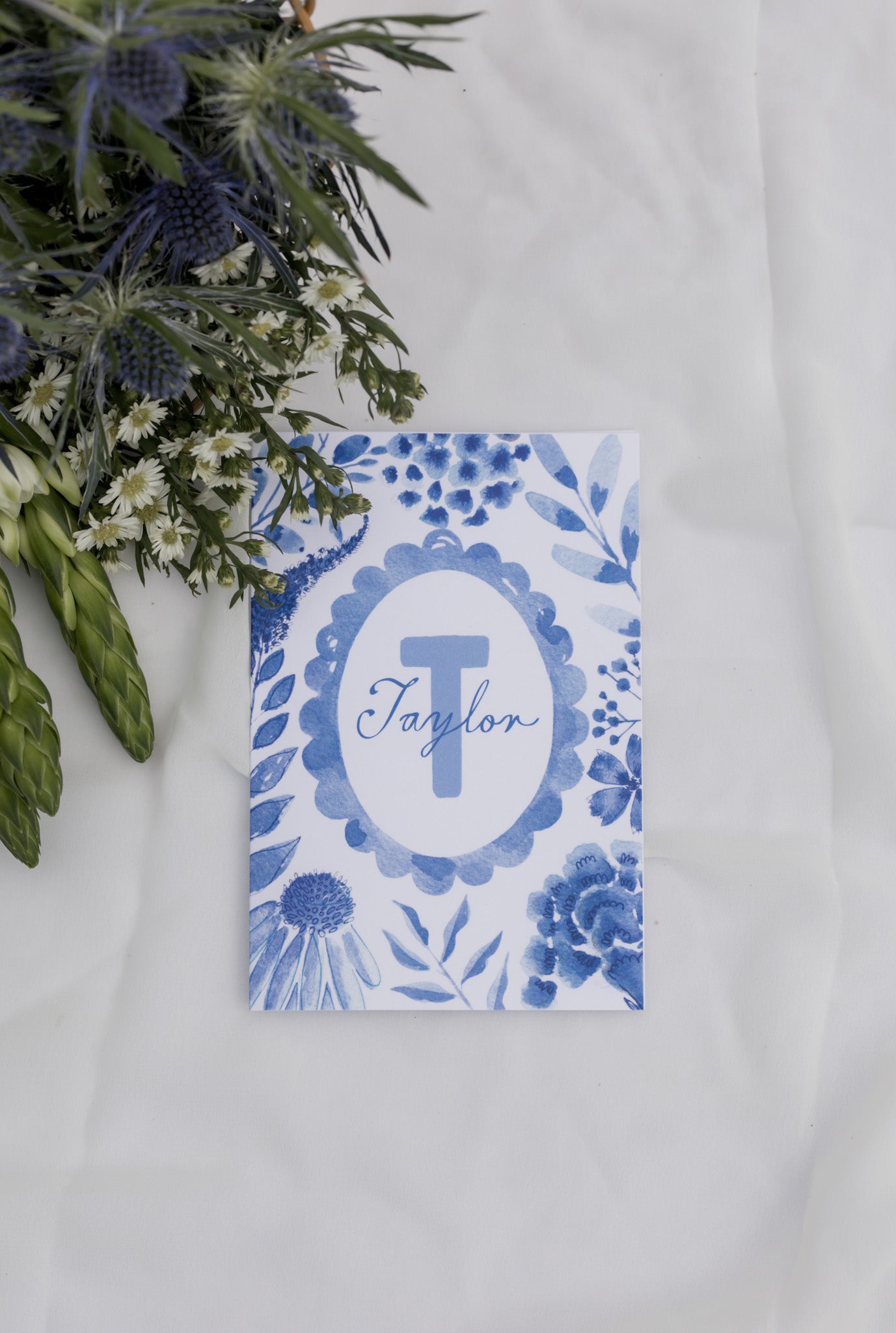 Personalized card with blue floral design and 'Taylor' monogram on a white surface with greenery.