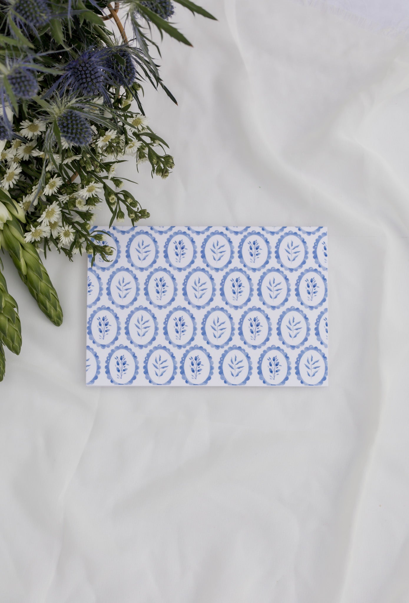 Card with blue floral pattern on a white surface with flowers in the background