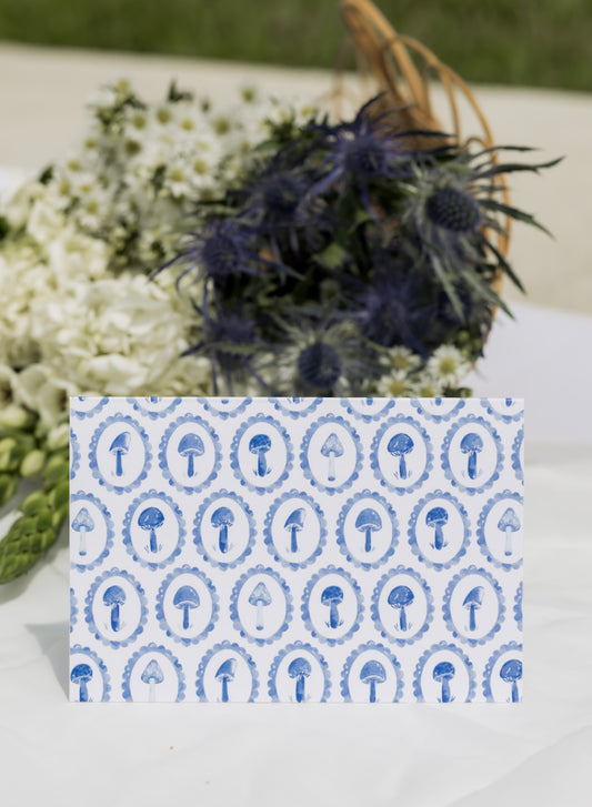Card with blue mushroom pattern on a white surface with flowers in the background