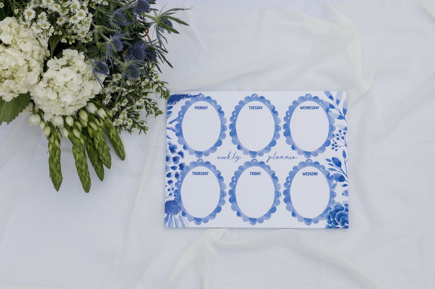 Weekly planner with blue floral design on a white surface with flowers.