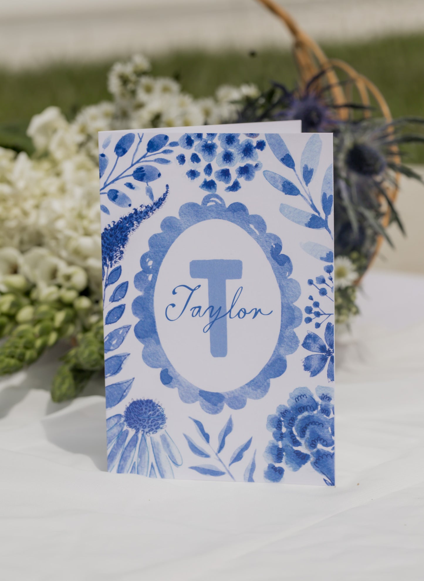 Personalized card with blue floral design and 'Taylor' monogram on a white surface with greenery.