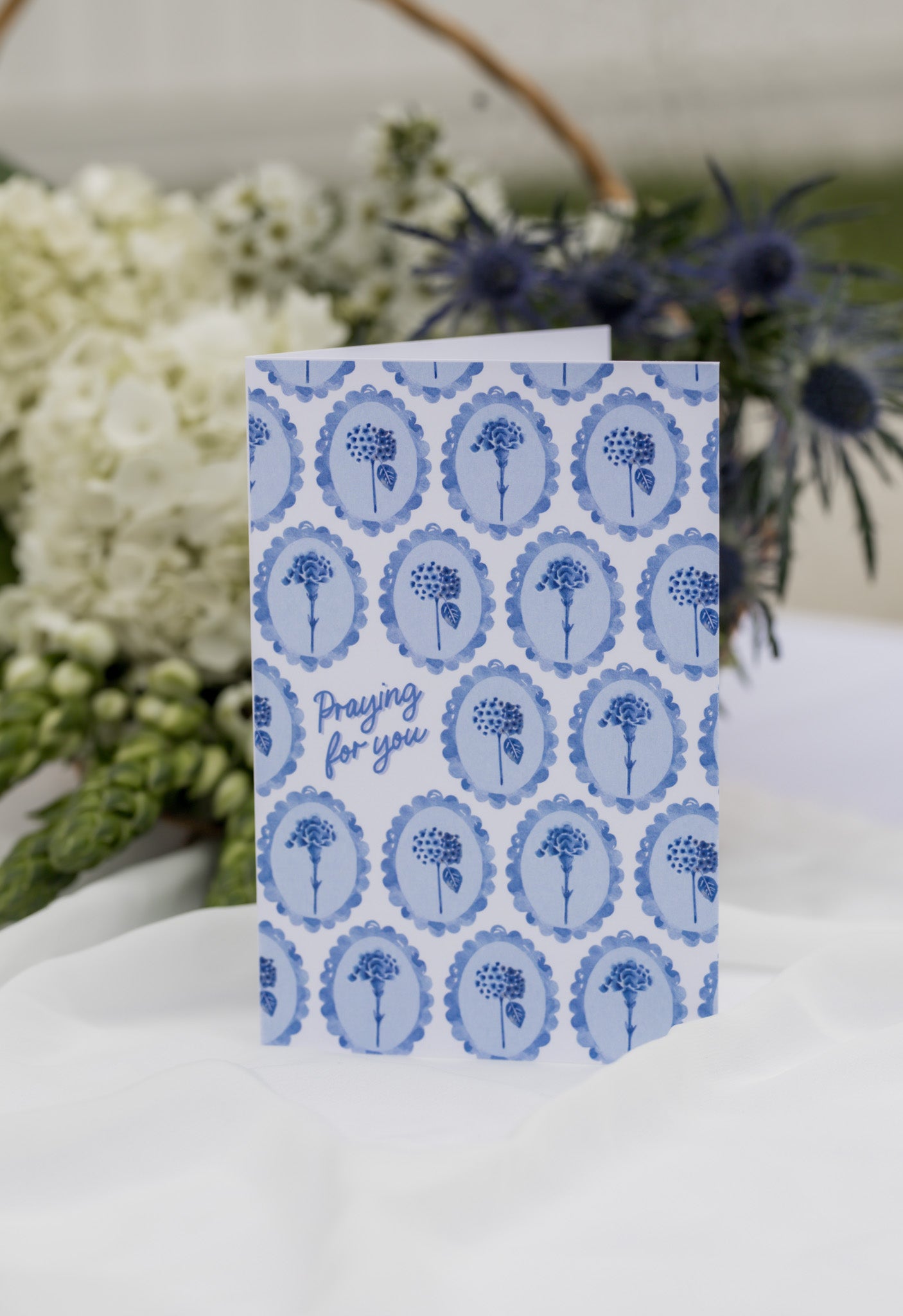 Blue patterned card with text 'Praying for you' on a white surface with flowers in the background