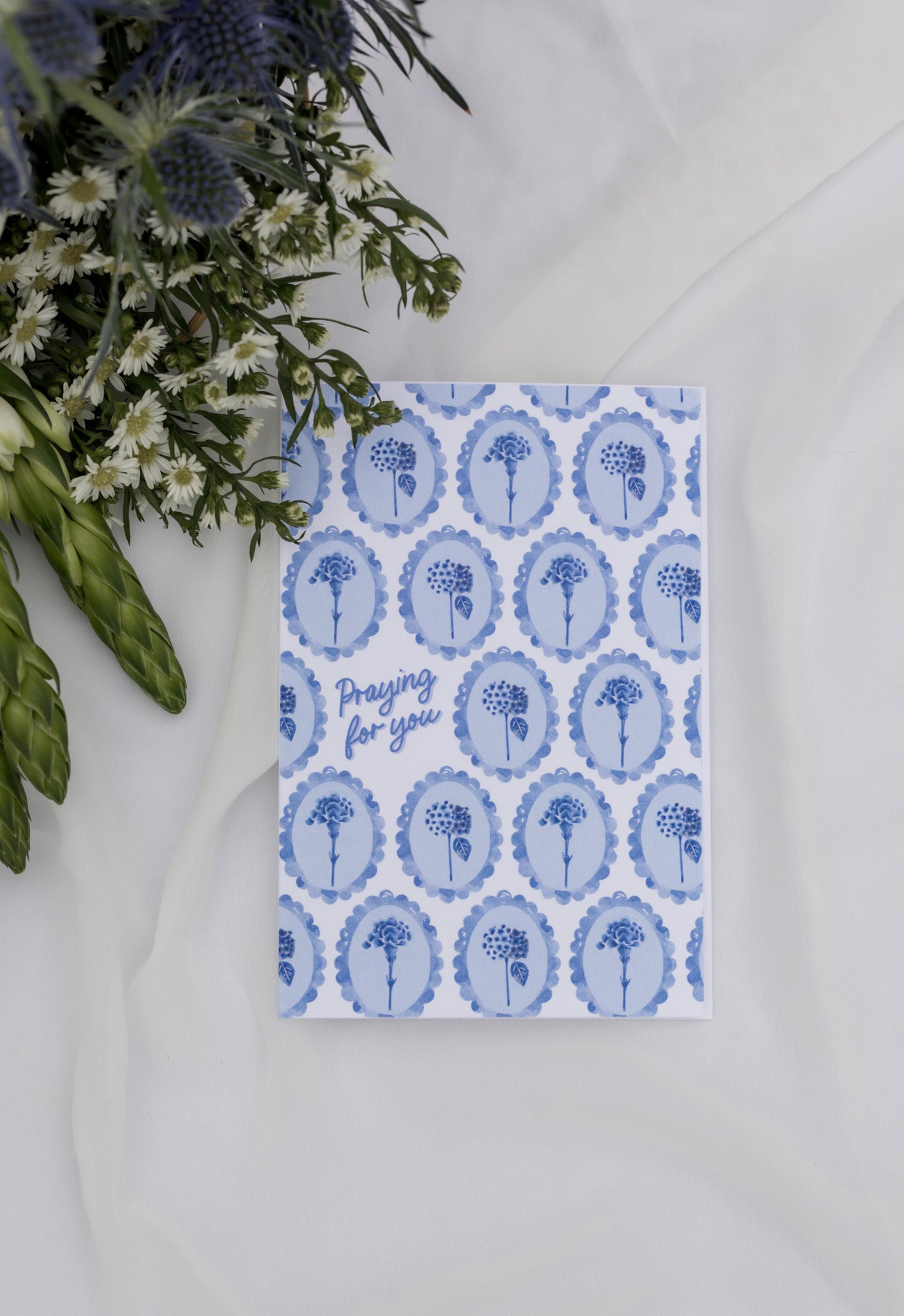Card with blue floral pattern and text 'Praying for you' on a white surface with flowers.