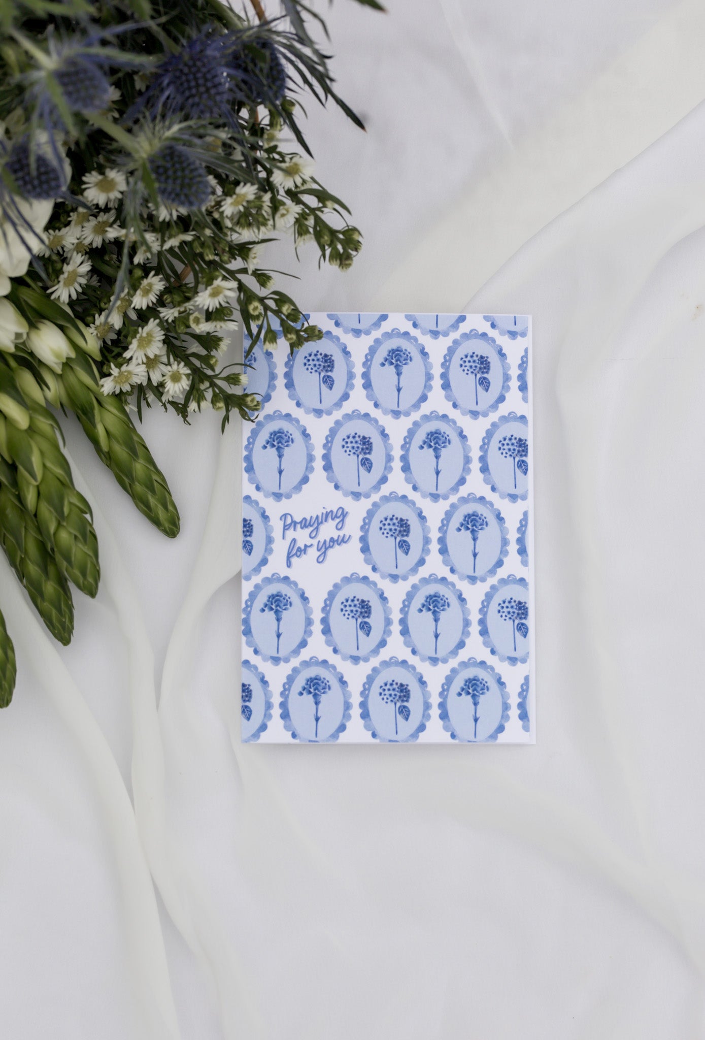 Card with blue floral pattern and text 'Praying for you' on a white surface with greenery.