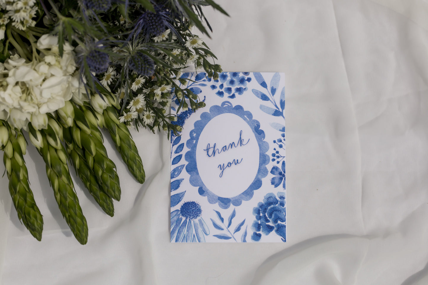 Blue floral card with 'thank you' text next to a bouquet of flowers on a white fabric background