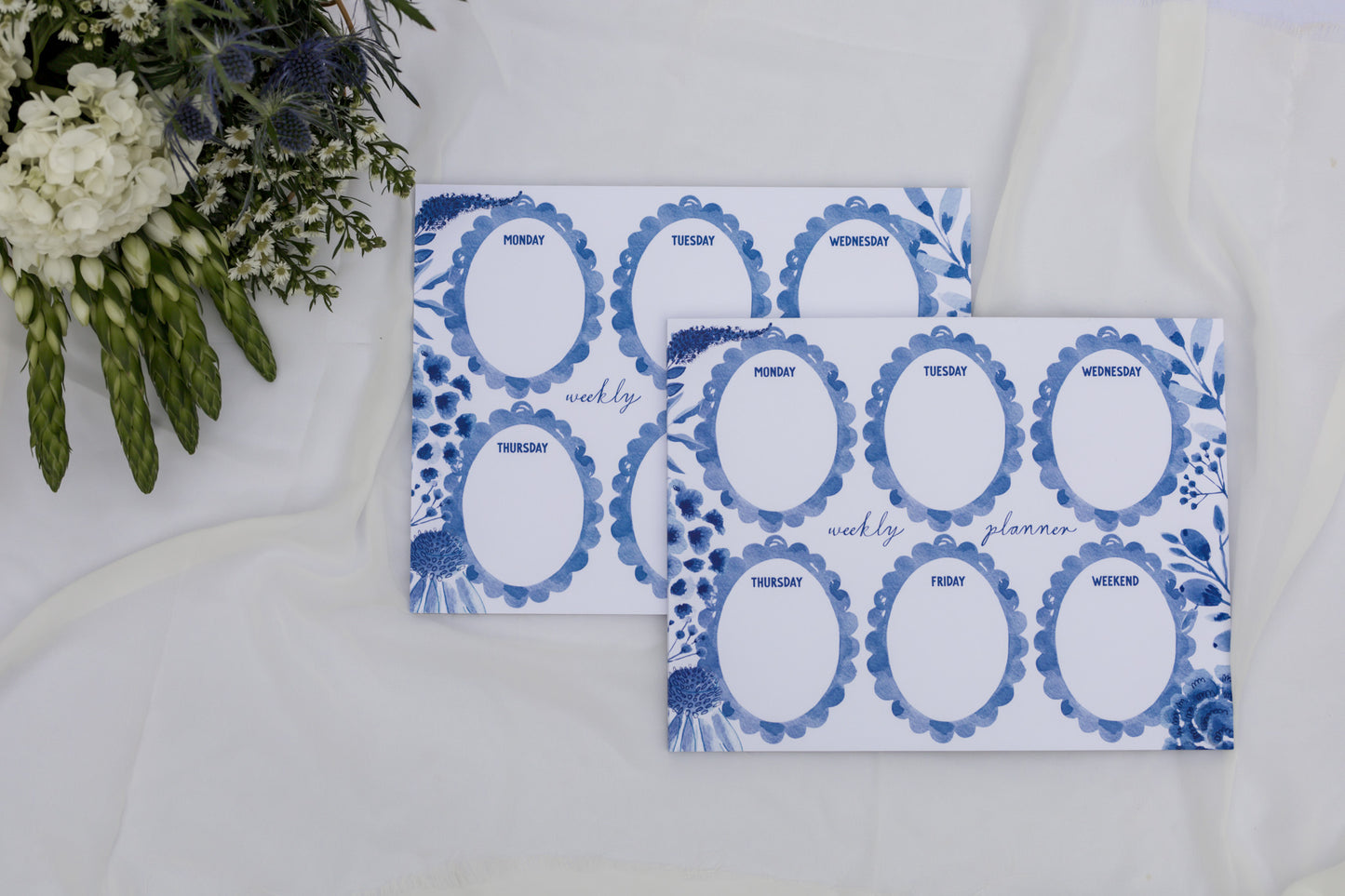 Two blue and white weekly planner sheets on a white surface with flowers in the background.