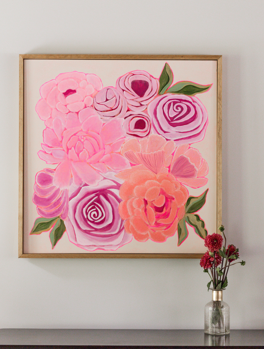 Framed wall art featuring a stylized floral bouquet in shades of pink, coral, and green, displayed on a light gray wall above a dark surface with a small vase of red flowers.