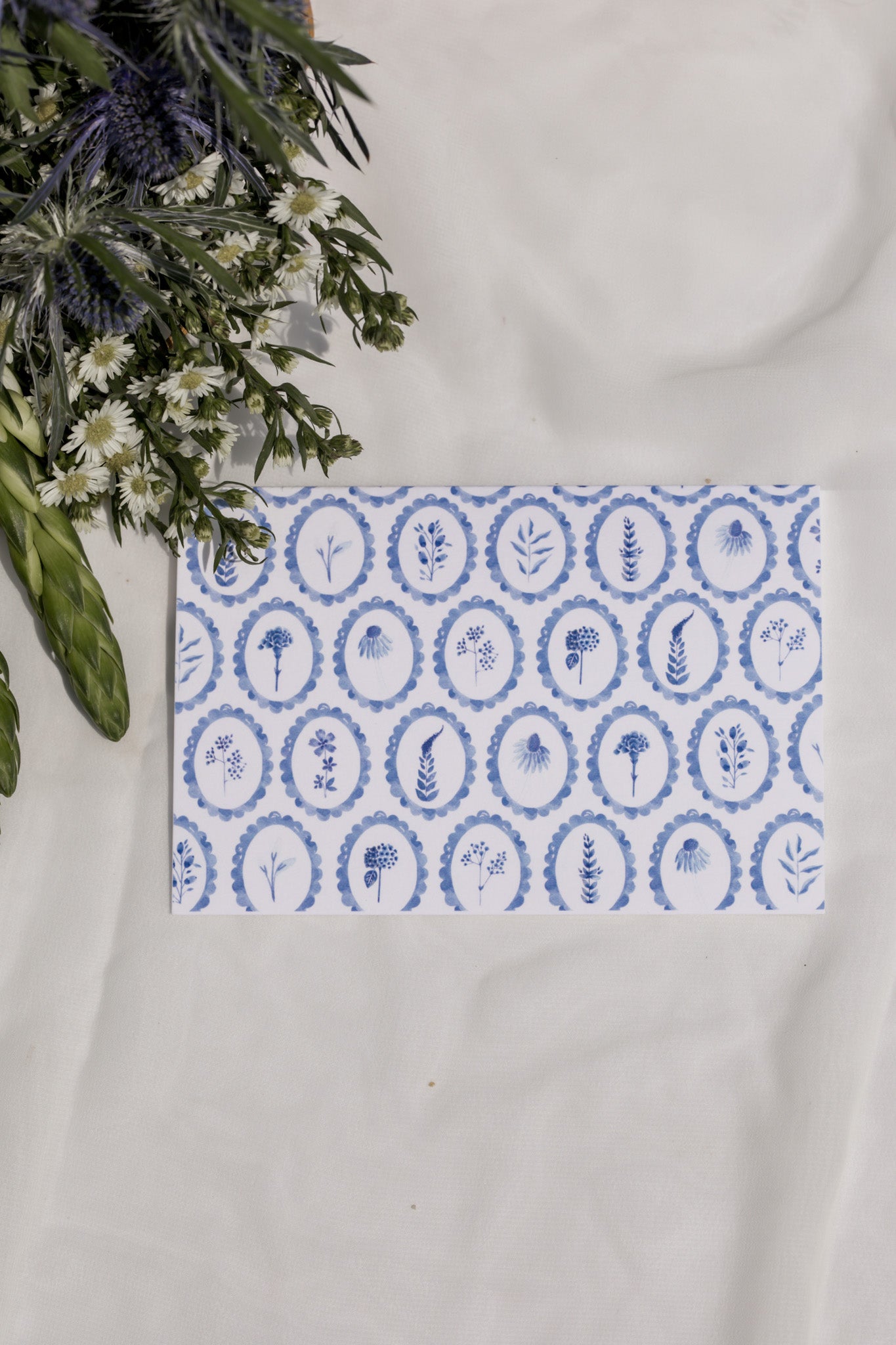 Decorative card with blue circular patterned wildflowers on a white surface with flowers in the background