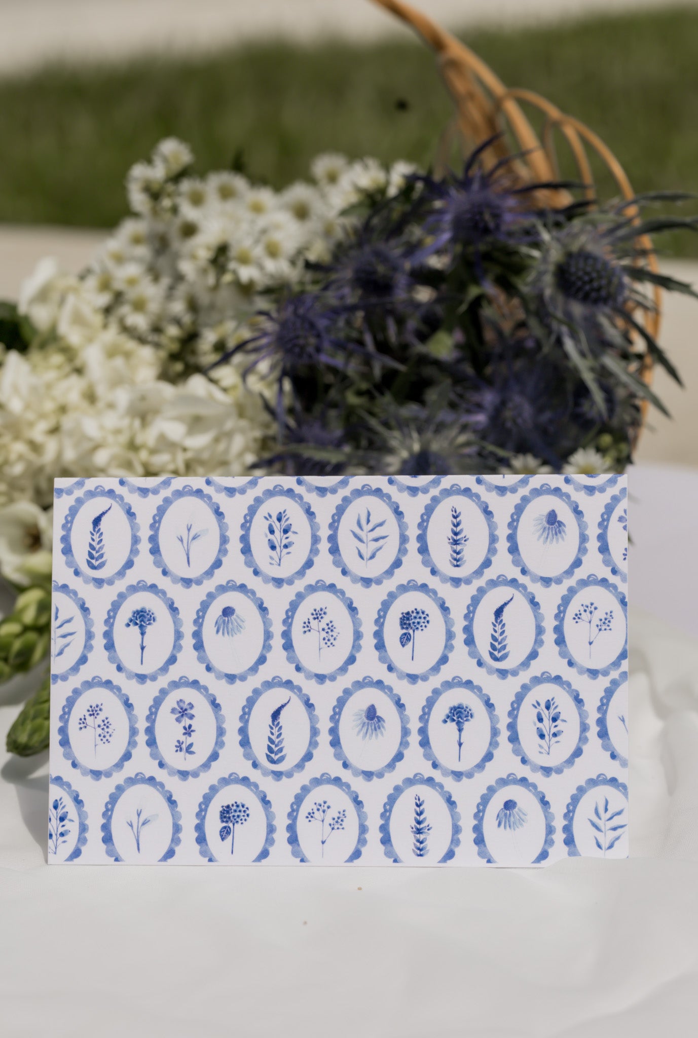 Decorative card with blue circular patterned wildflowers on a white surface with flowers in the background