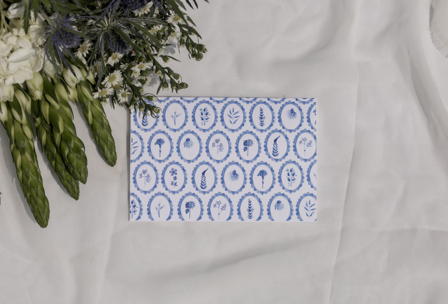 Decorative card with blue circular patterned wildflowers on a white surface with flowers in the background