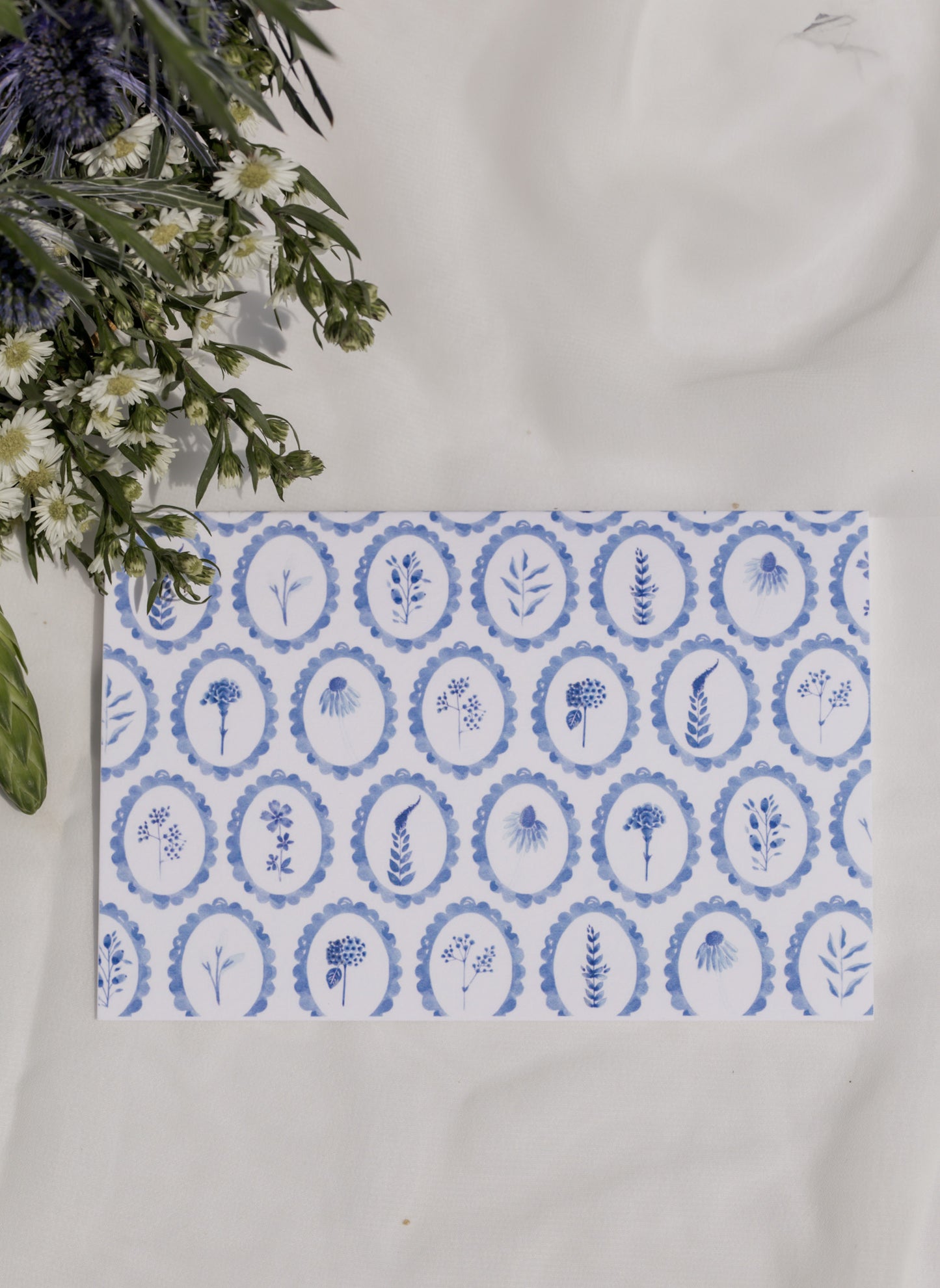 Decorative card with blue circular patterned wildflowers on a white surface with flowers in the background